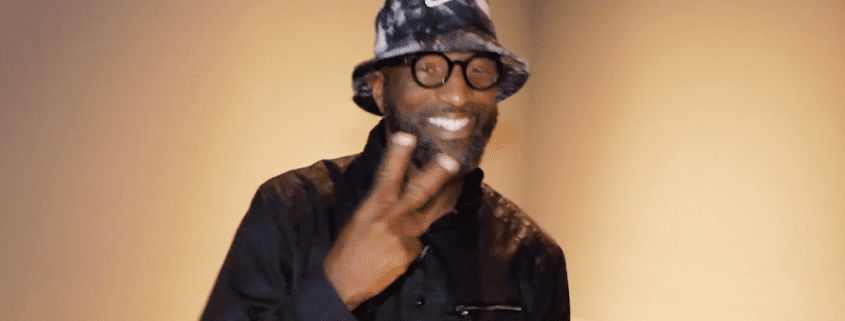 Smiling man wearing hat, flashing peace sign.